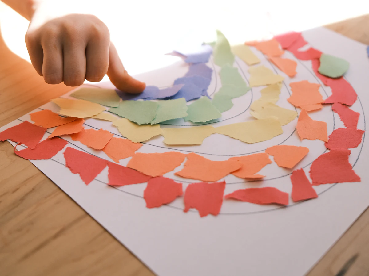 A child gluing colourful paper scraps onto a rainbow drawing