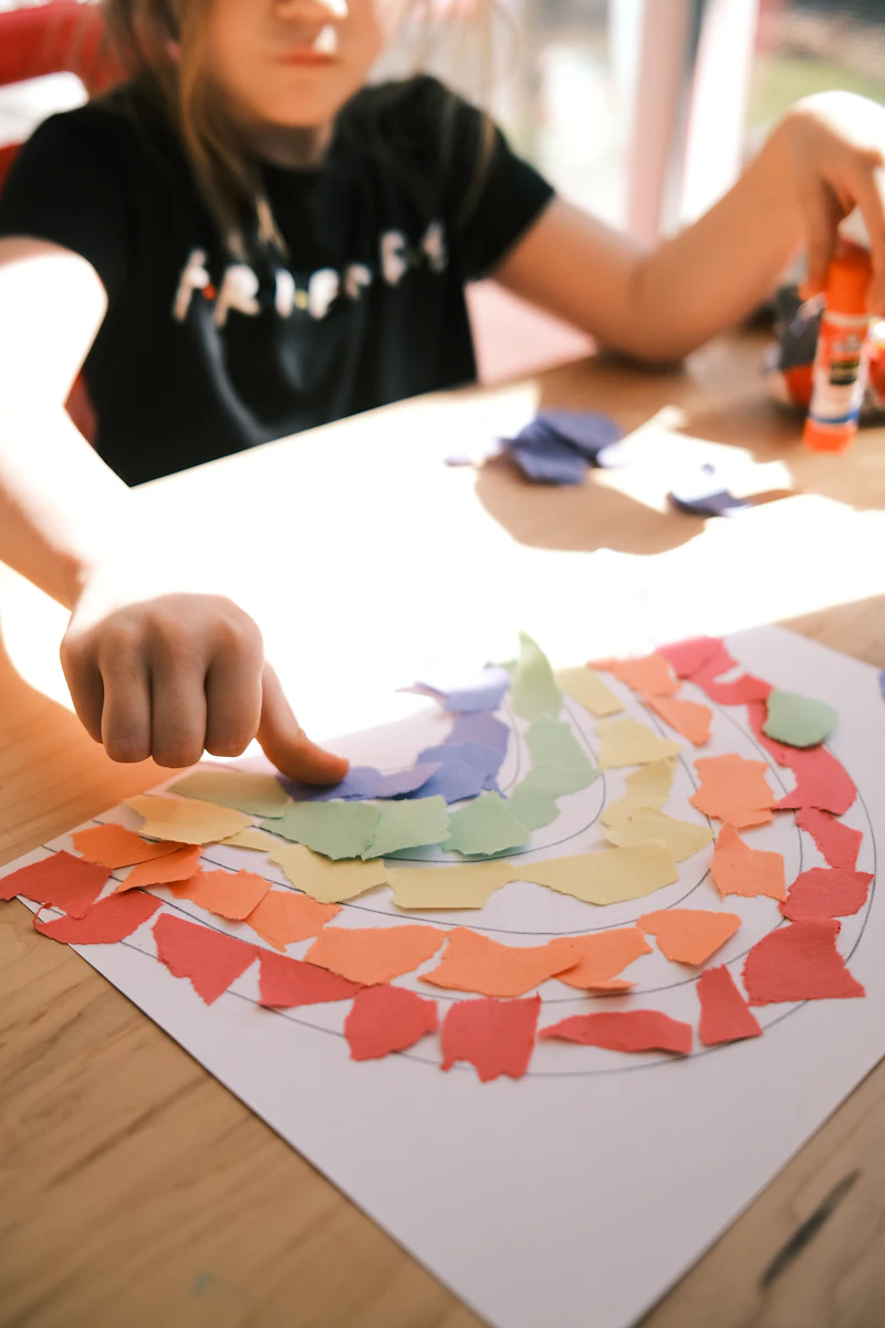 A child gluing coloured paper scraps onto a rainbow